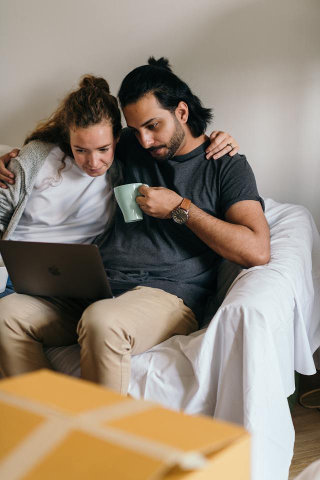 A young couple on the couch looking at a laptop. This image represents two buyers searching for a home or real estate agent in the Albuquerque area.