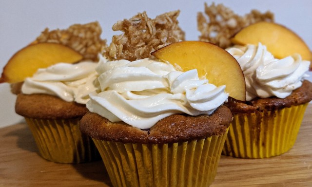 group of three baked and frosted nectarine cupcakes. Whipped, white and fluffy frosting a slice of fruit and oat crisp on top